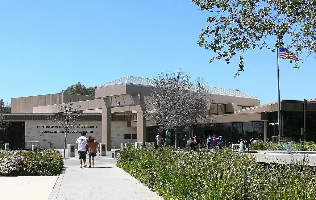 The entrance to Huntington Beach Central Library, a low modernist building with people walking the path in through landscaped native plantings.